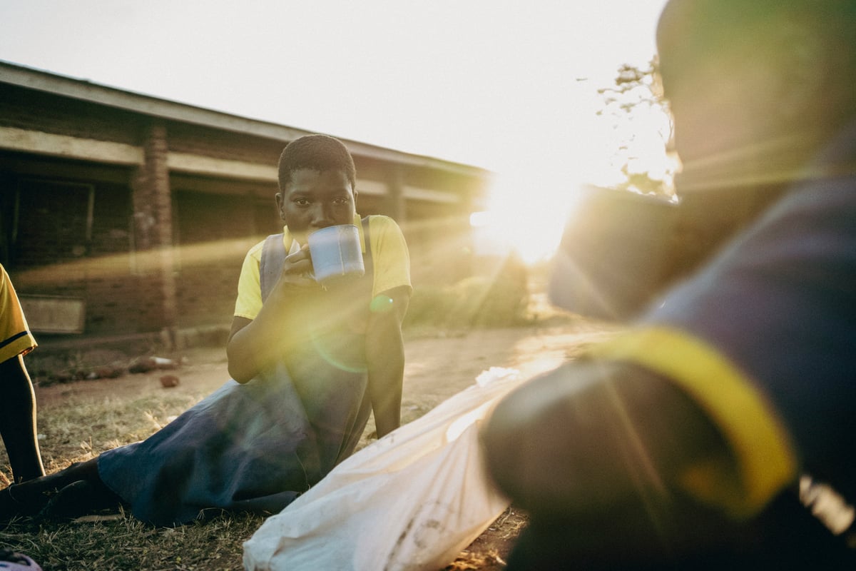 Una estudiante con uniforme amarillo bebe de una taza al atardecer, con el sol de frente.