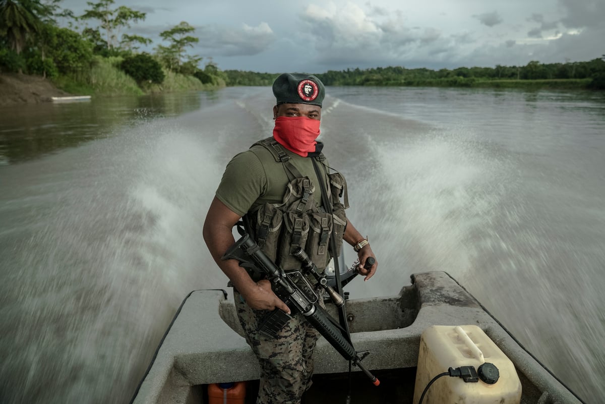 Un hombre armado, con boina y el rostro cubierto por un pañuelo rojo, de pie en la popa de una lancha que abre una estela en un río.