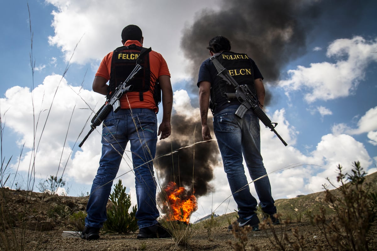 Dos agentes armados, de espaldas, observan una columna de humo negro y fuego en un campo abierto.