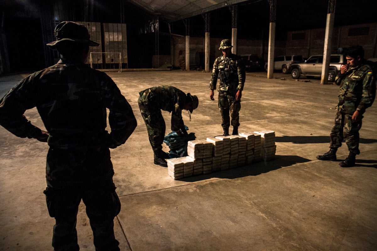 De noche, en una nave, varios militares rodean paquetes apilados en el suelo.
