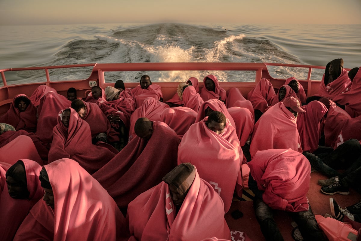 Personas rescatadas, envueltas en mantas rojas, en la cubierta de popa de un barco de salvamento. Detrás, la estela sobre el mar.